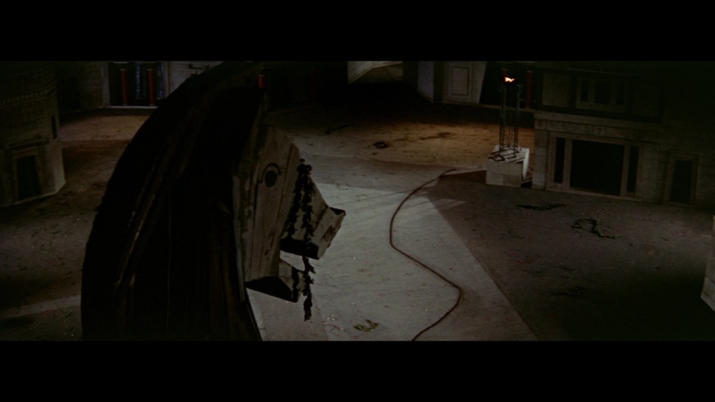 A large wooden horse structure in an empty, dimly lit indoor space, with scattered debris and a winding cable on the floor.