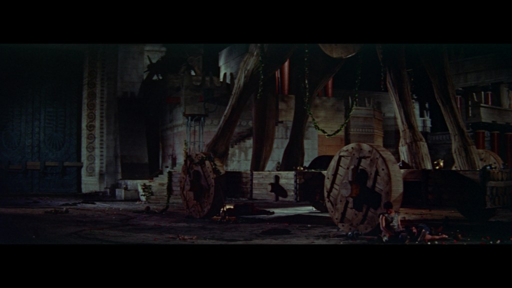 Dimly lit scene featuring a large wooden structure resembling a trojan horse, surrounded by ancient architecture and two figures sitting on the ground.