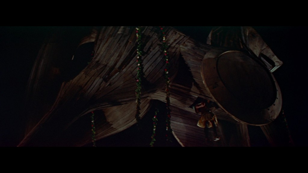 A large wooden structure resembling a colossal figure, shrouded in darkness, with a person hanging from it, adorned with garlands of flowers.