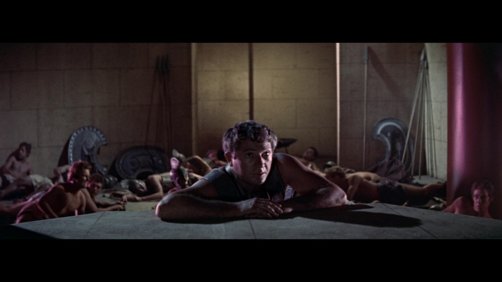 A man resting on a stone surface, gazing intently, while multiple figures lie in the background, partially visible, against a dimly lit stone wall.