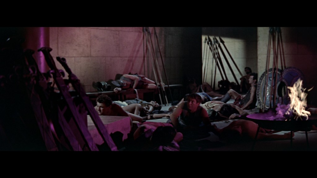 A dimly lit room with several individuals lying on the ground, some are resting while others are seated. In the foreground, there are several swords and shields arranged, and a small fire burning in a pot. The atmosphere suggests a historical or dramatic setting.