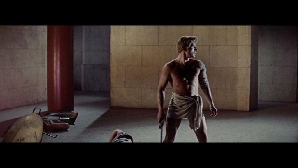 A muscular man standing shirtless in a large, empty room, holding a weapon and looking around cautiously. The background features concrete walls and a few scattered items on the floor.
