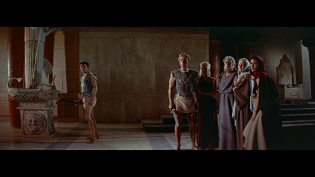 A group of ancient Greek figures stands in a dimly lit temple, with one male figure in the foreground holding a sword. The others appear to be in conversation, dressed in traditional robes.