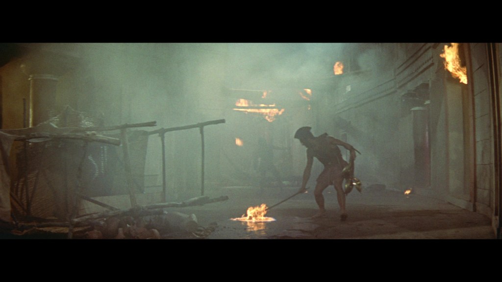A dimly lit scene with smoke and flames, depicting a person in ancient attire tending to a fire in a deteriorating environment.