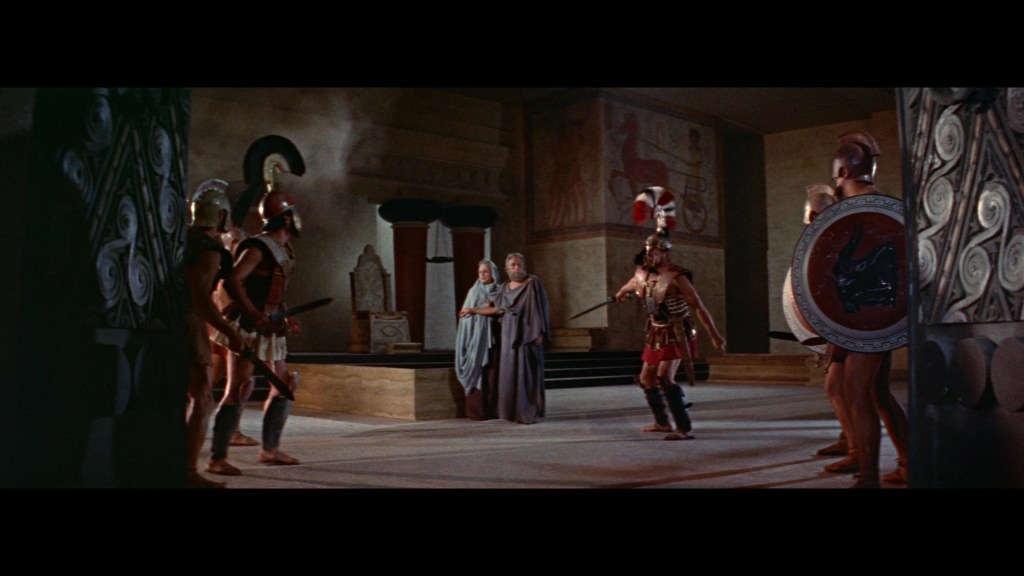 A scene depicting ancient warriors in armor and helmets engaged in a tense standoff in a historically styled interior, with two cloaked figures observing the confrontation.