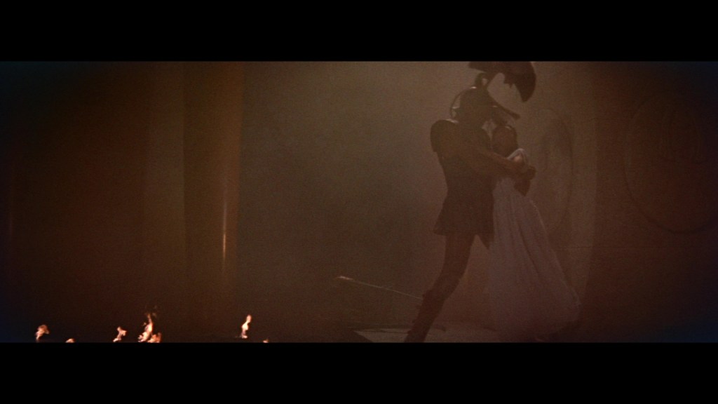 A scene depicting a warrior in armor holding a woman in a flowing dress, amidst a smoky environment with faint flames in the foreground.