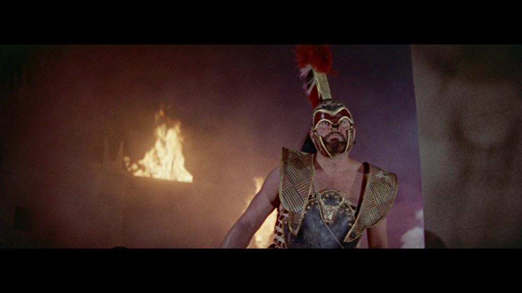 A muscular warrior in ornate armor and a plumed helmet stands amidst smoke and fire, exuding a fierce presence.