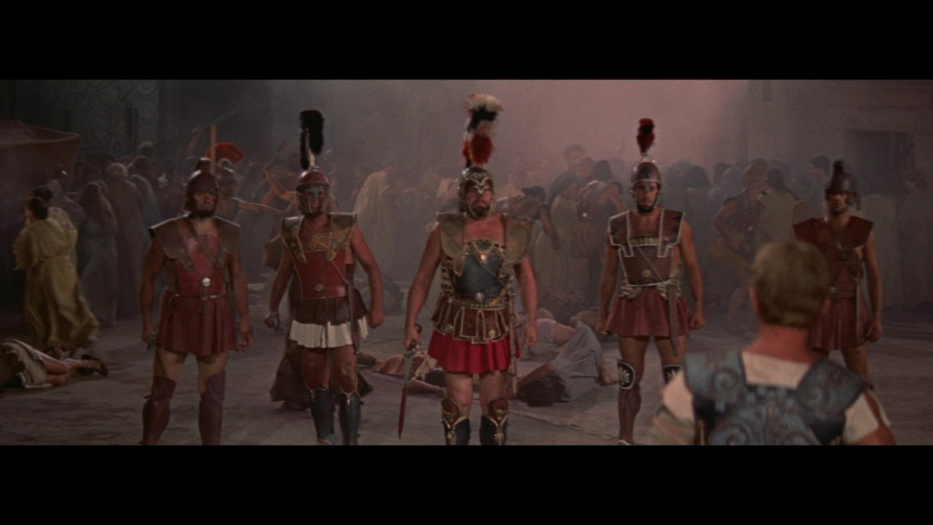A group of Roman soldiers in armor and helmets stands in a dramatic scene, with a blurry crowd and scattered debris in the background.