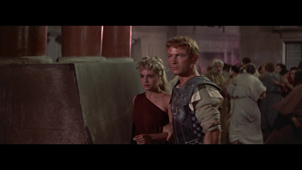 A scene depicting two characters in a historical or ancient setting, with the male character wearing armor and the female character in a brown dress. They appear to be in a crowded environment, suggesting an atmosphere of tension or excitement.