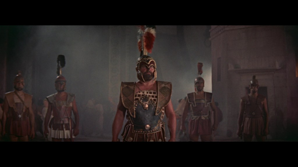 A group of ancient warriors in armor and helmets stands in a dark, misty setting, with a prominent leader in the foreground wearing ornate armor and a plume on their helmet.