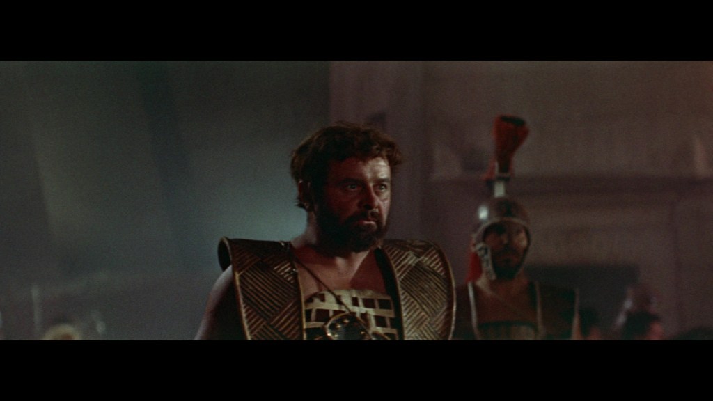 A scene featuring a bearded man in ancient armor, with a serious expression, standing in a dimly lit setting alongside another soldier in a helmet.