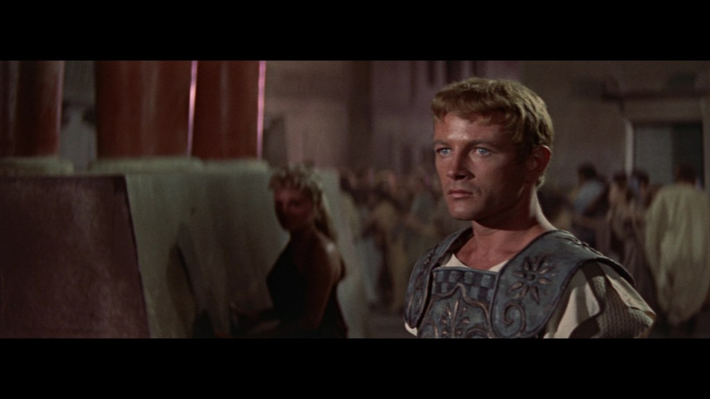 A man in ancient Roman attire with a serious expression stands in a crowded setting, surrounded by pillars and other figures in historical clothing.