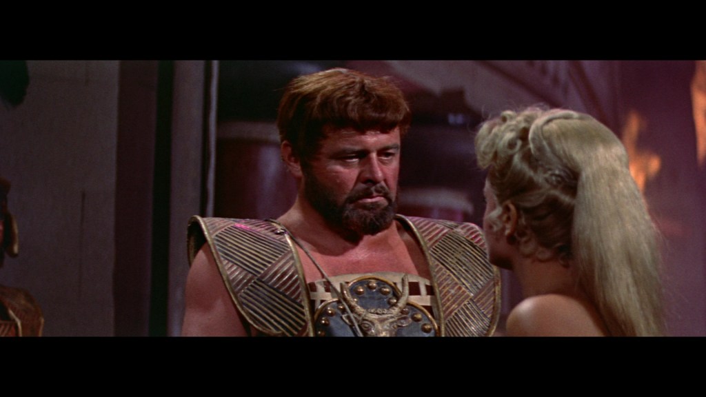 A man in ancient-style armor with a beard and intense expression, engaging in conversation with a woman with long, blonde hair, in a historical setting.