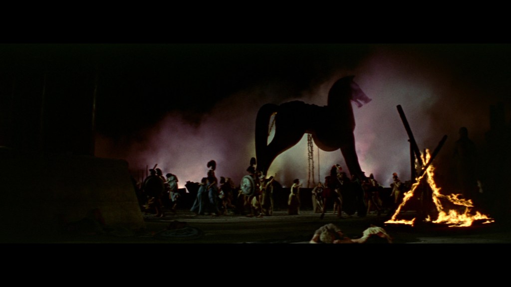 A large silhouette of a wooden horse dominates the scene, surrounded by a crowd of ancient warriors. Smoke and fire illuminate the dark background, creating a dramatic atmosphere.