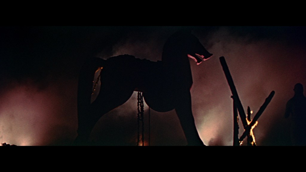 Silhouette of a large wooden horse against a smoky background, with faint figures visible nearby.