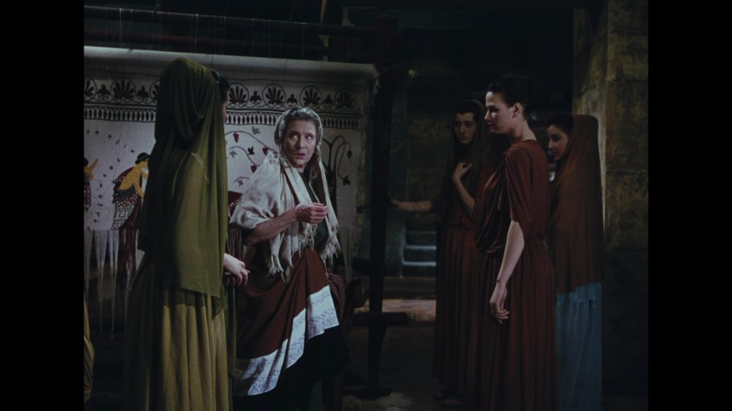 A scene featuring a group of women in ancient attire, engaged in conversation. An older woman gestures as she addresses the others, who are standing nearby in a dimly lit room with a tapestry in the background.