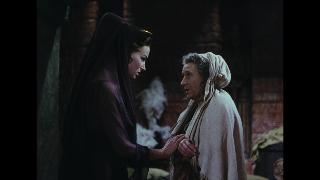 A scene depicting two women engaged in a heartfelt conversation in a dramatic setting. One woman is wearing a dark veil, while the other is dressed in a light-colored shawl, both displaying expressions of emotion.