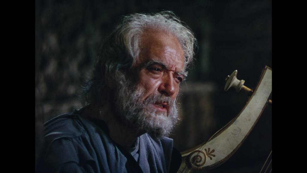 An elderly man with long gray hair and a beard appears emotionally intense, sitting near a stringed instrument in a dimly lit environment.