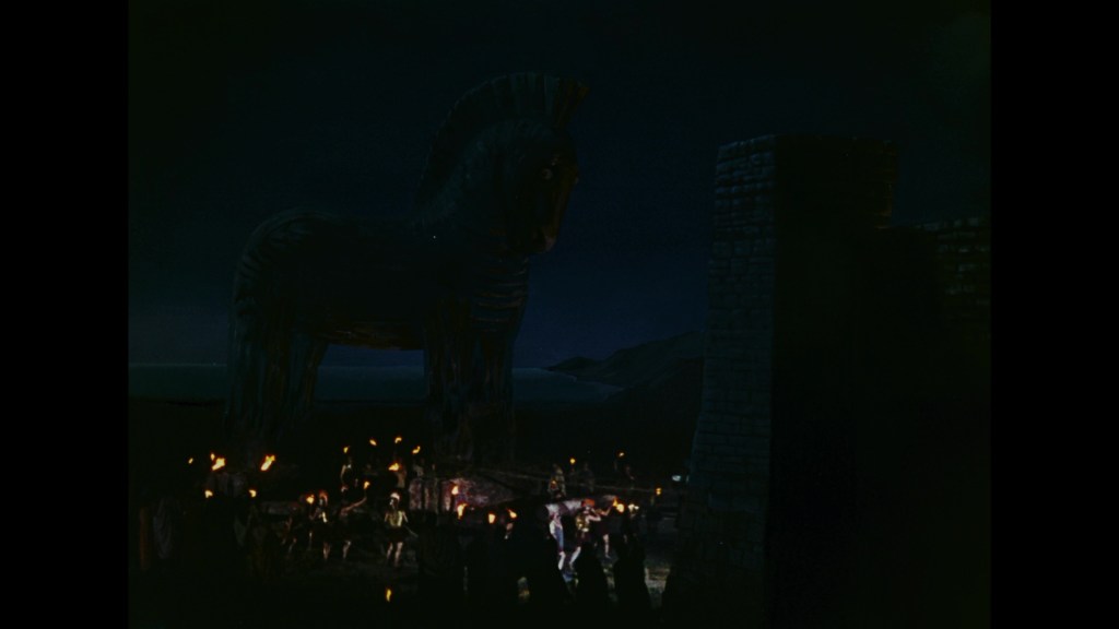 A large wooden Trojan horse stands prominently at night, surrounded by people holding torches near a walled city.
