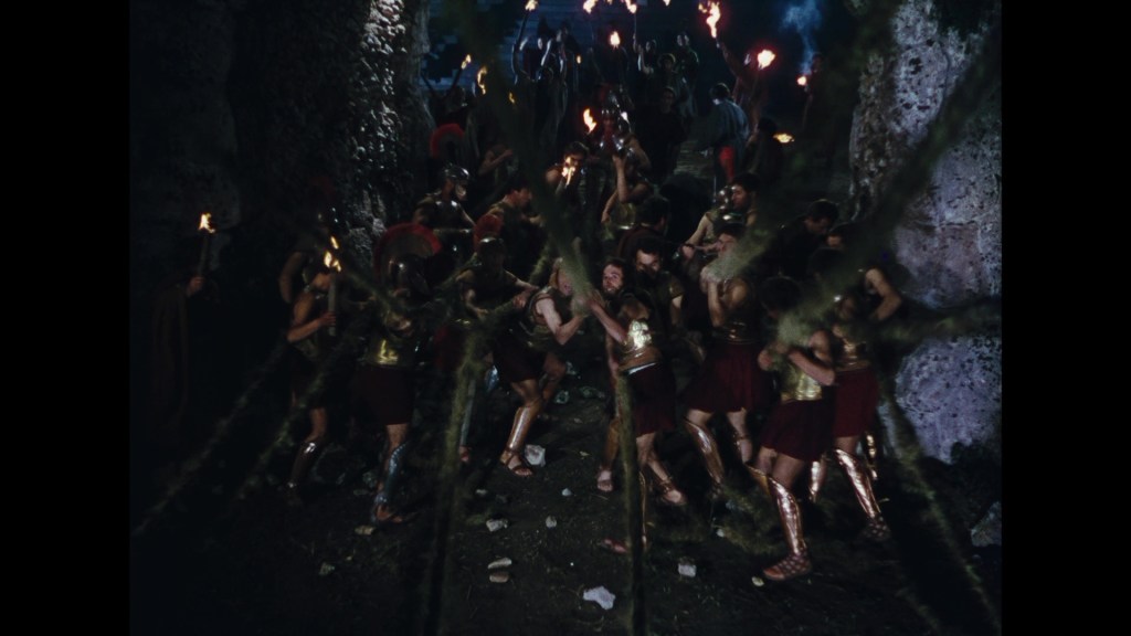 A group of ancient warriors in bronze armor and gladiator-like attire are gathered in a rocky cave, holding torches and appearing to be in a tense situation. Some are pulling on ropes, while others look on, set against a dark background illuminated by the torchlight.