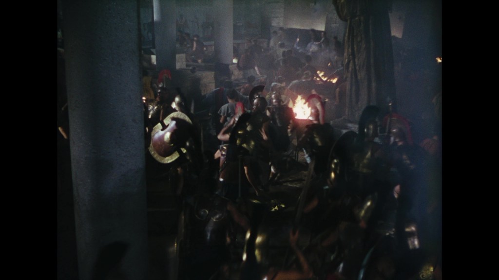 A chaotic scene featuring armored warriors in a dark, smoky environment, engaged in battle with weapons and shields raised, depicting an ancient combat scenario.