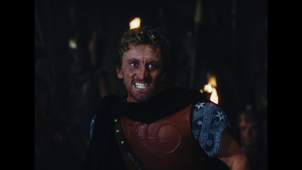 A man with curly hair, wearing a leather tunic and cape, exhibits an intense expression in a dimly lit setting filled with shadows and torches.