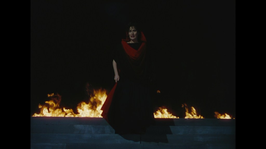 A dramatic scene with a woman dressed in a dark gown and a red shawl standing in front of flames against a black backdrop.