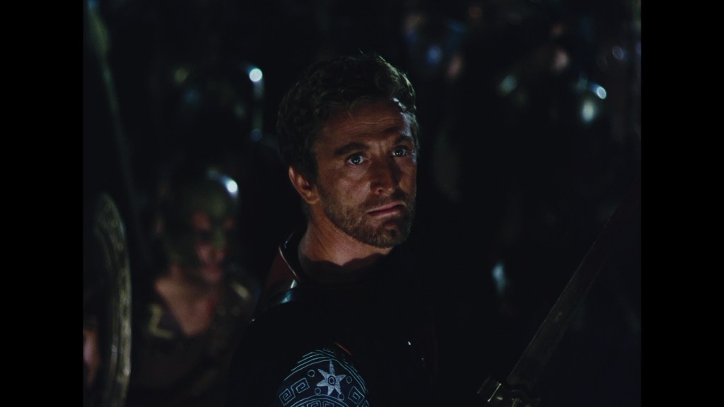 A solemn man with short, tousled hair and a serious expression, wearing armor and holding a sword, stands in a dimly lit environment surrounded by figures in the background.