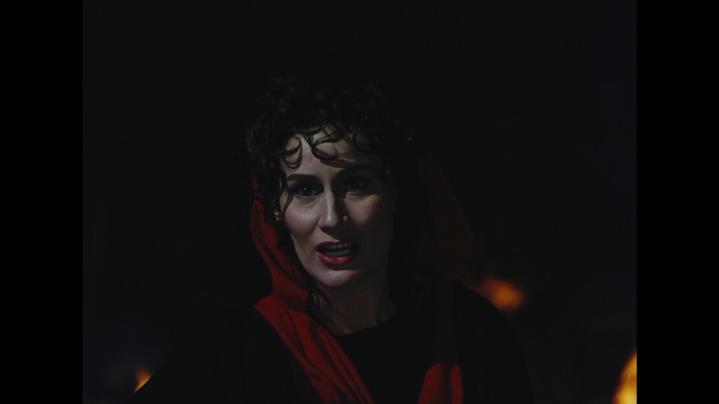 A woman with curly dark hair wearing a black outfit and a red hood stands against a dark background, speaking with an intense expression.