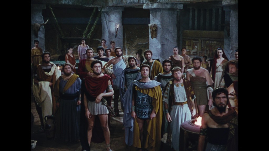 A group of men dressed in ancient Greek clothing standing in a dimly lit stone chamber, with some wearing togas and others in armor, all facing forward.