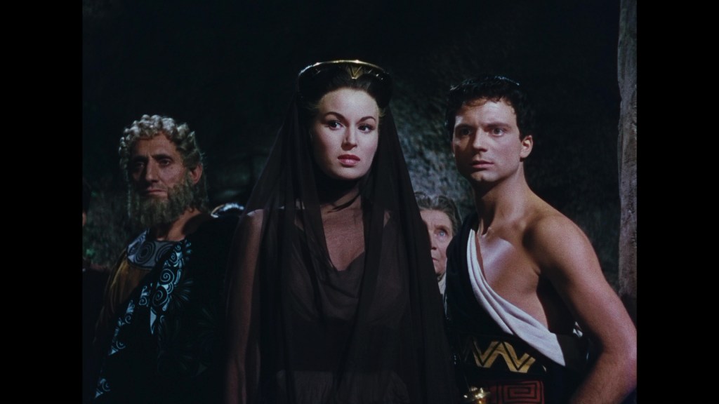 A group of four individuals standing closely together in a dimly lit scene, with a central figure in a dark veil, flanked by two men in historical attire, one with a beard and another dressed in a partially exposed costume.
