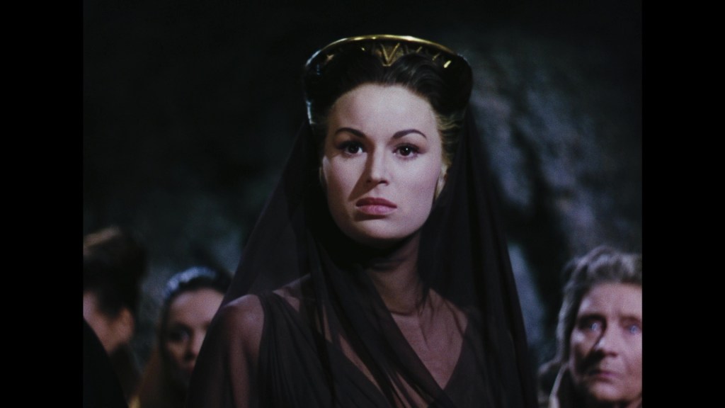 A woman in a dark veil and elegant gown, striking a serious pose, surrounded by other figures in a dimly lit setting.