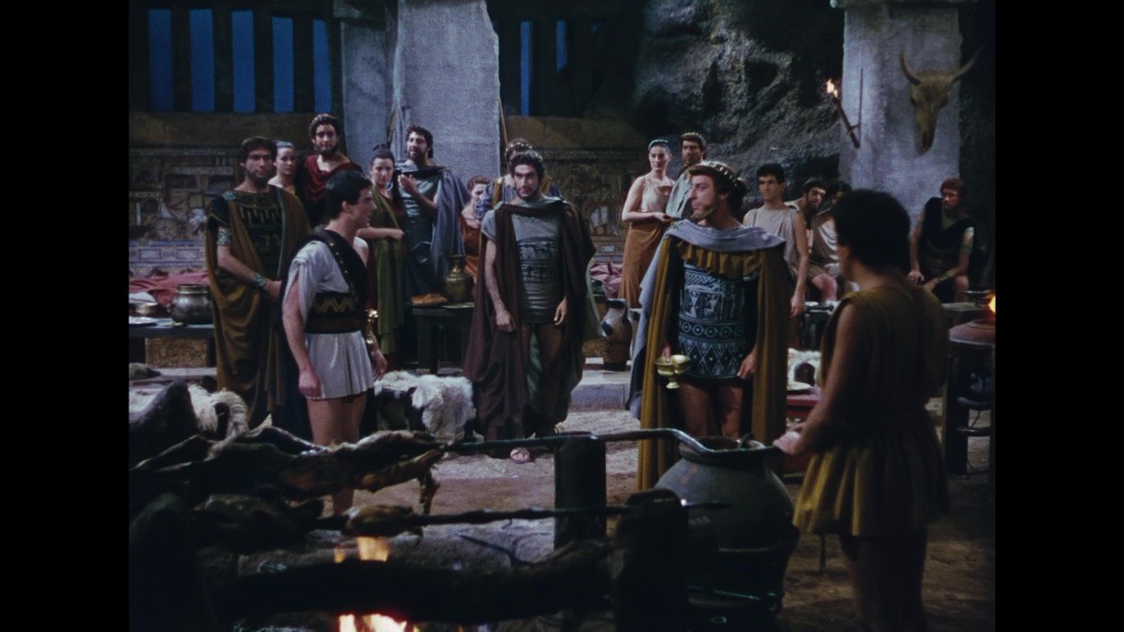 Ancient Greek gathering in a cave-like setting, featuring men and women dressed in traditional robes and tunics, engaged in conversation.