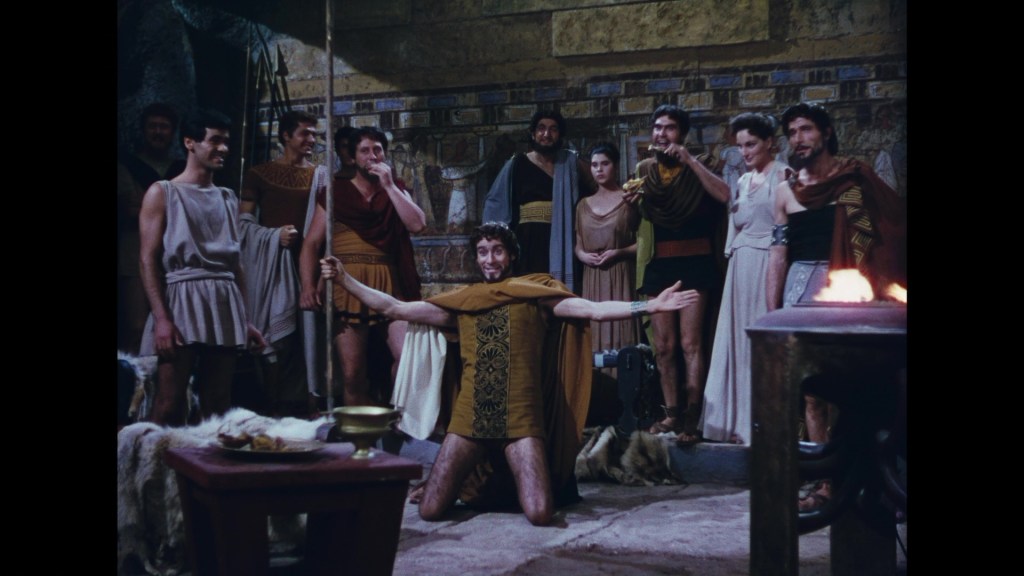 A group of ancient Greek characters dressed in traditional attire, gathered around a central figure kneeling and gesturing dramatically. The setting appears to be in a stone chamber with ancient decorations in the background.
