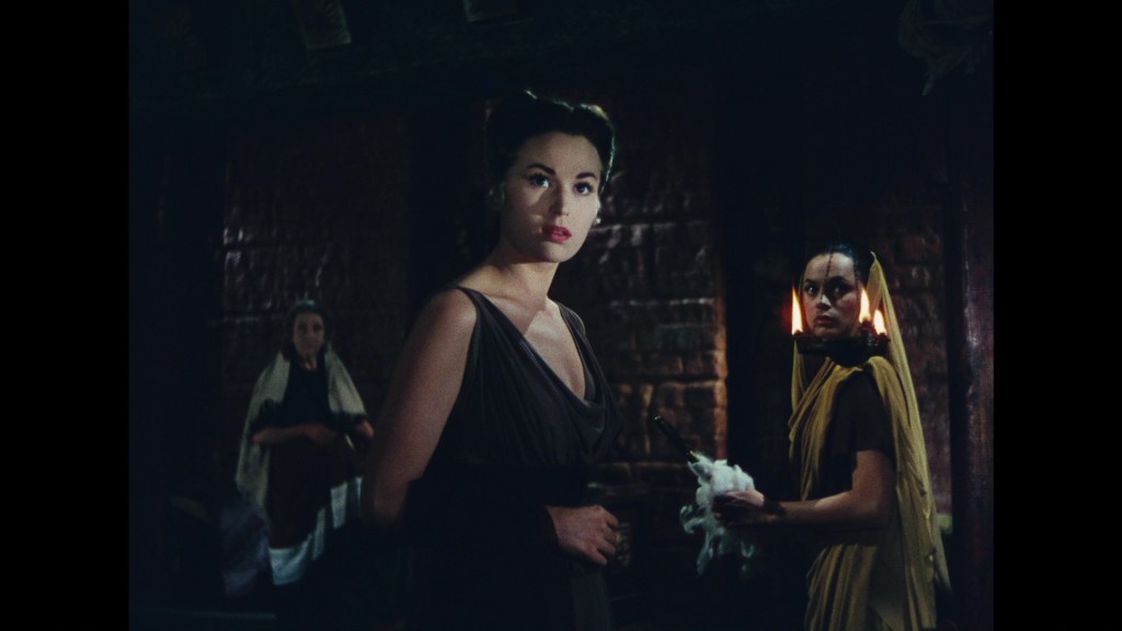 A dramatic scene featuring three women in a dimly lit room, with one woman in the foreground looking directly at the viewer while the other two appear engaged in different activities.