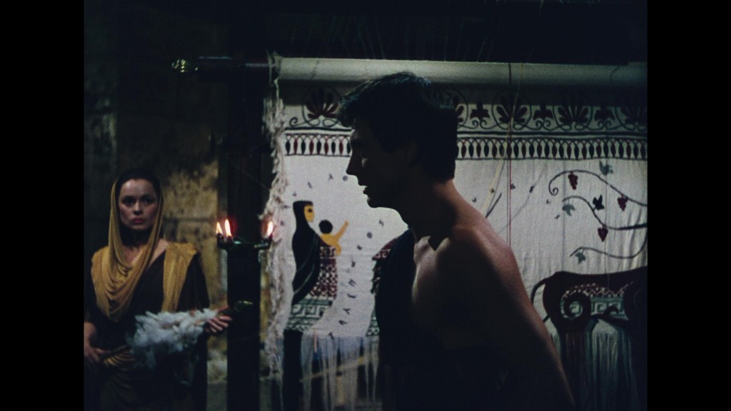 A dramatic scene featuring a woman in traditional attire holding feathers, watching a shirtless man in a dimly lit room with intricate wall decorations.