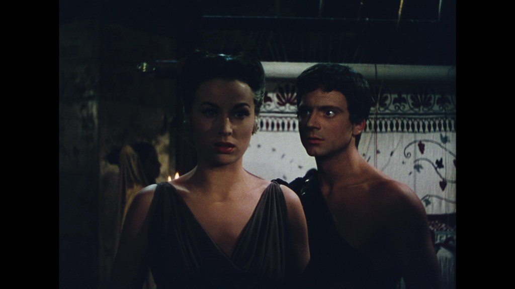 A serious-looking woman in a dark dress stands beside a young man in a sleeveless shirt, with an ornate backdrop featuring decorative patterns.