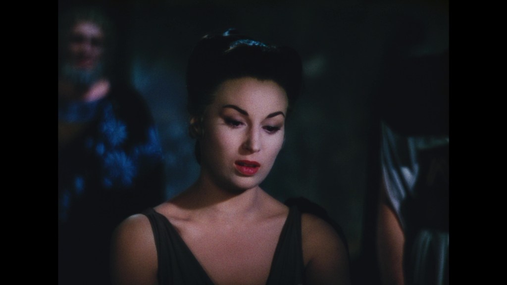 A woman in a dark setting, featuring dramatic lighting and expressions, showcasing a contemplative mood. She has dark hair styled elegantly and is wearing a sleeveless dress.