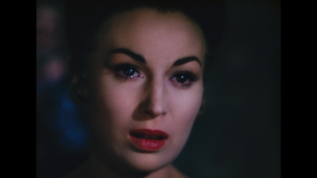 Close-up portrait of a woman with dark hair, bold eyebrows, and red lips, showing a serious expression against a blurred dark background.