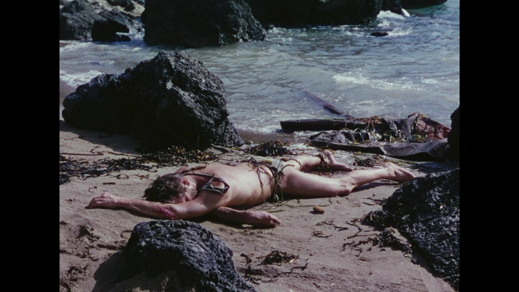 A person lying on a sandy beach surrounded by rocks and seaweed, with waves in the background.