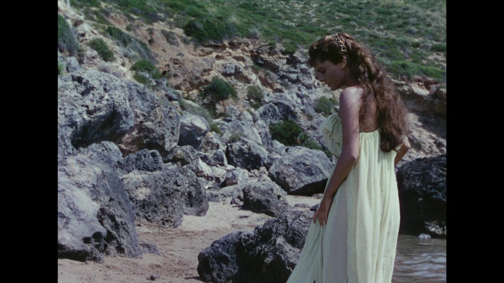 A woman in a flowing yellow dress stands on a rocky beach, gazing down at the ground.