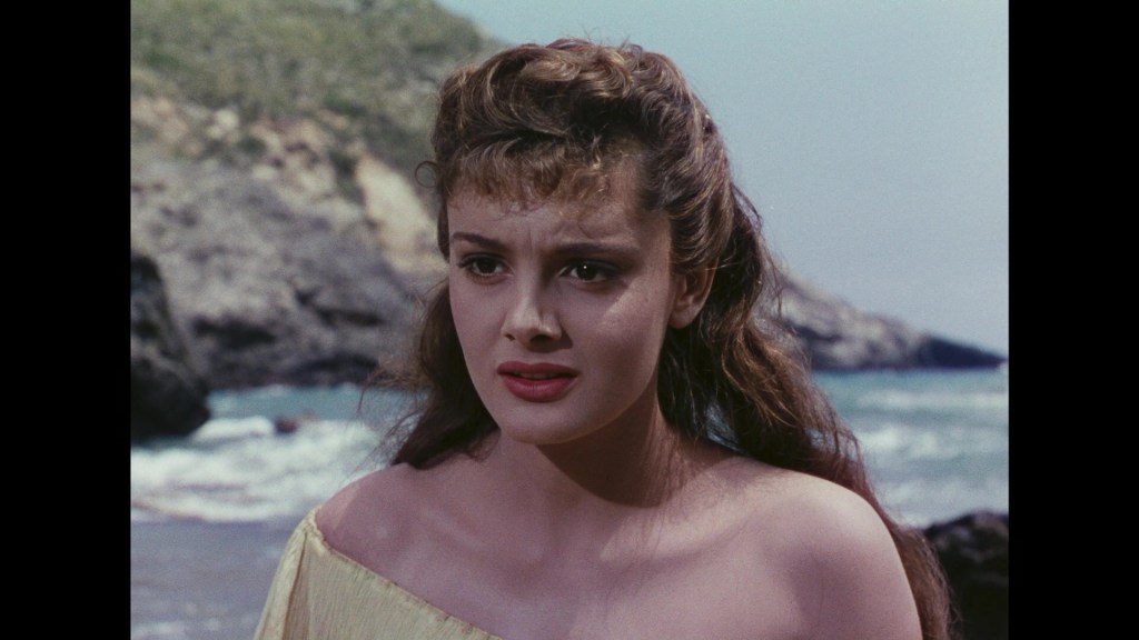 A young woman with long, wavy hair and a concerned expression stands by the ocean, wearing a strapless yellow dress.