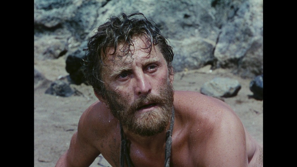 A close-up of a rugged man with a beard, looking distressed while sitting on a beach, with rocks and sand in the background.