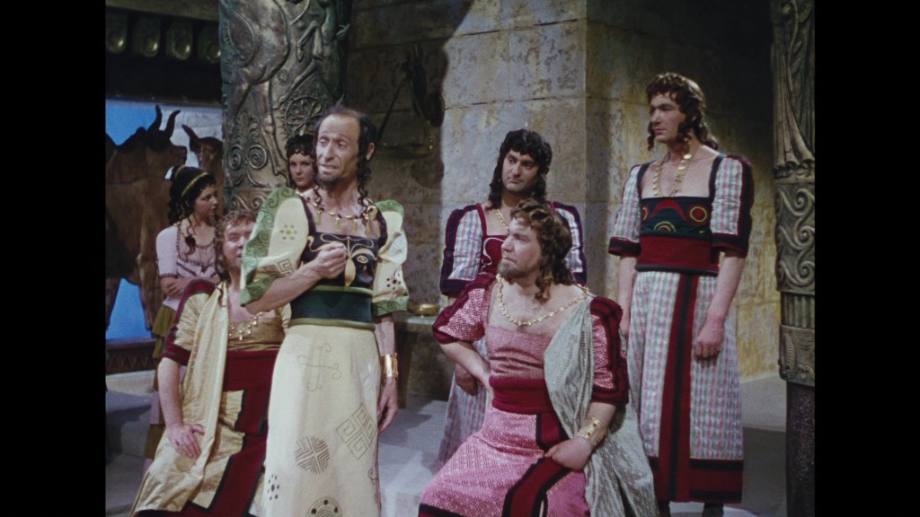 A group of actors dressed in ancient Greek-style costumes, engaged in a dramatic scene. The setting features large stone columns and decorative details, suggesting a historical or mythological context.