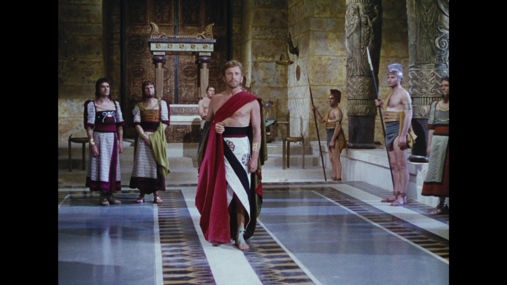A scene depicting several individuals in ancient attire, with one man in a prominent position wearing a red and black draped outfit, standing before an audience in a grand architectural setting.