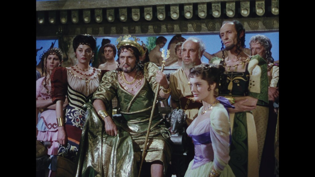 A group of people dressed in ancient Roman-style costumes, with a central figure wearing a regal outfit and a crown, seated on a throne. The scene includes men and women in elaborate attire, showcasing a dramatic expression and setting.