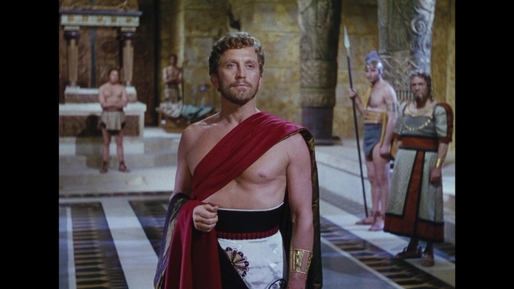 A male character in ancient attire stands confidently in a grand indoor setting, adorned with ornate architecture. He wears a red draped garment and a white garment with a decorative belt. Behind him are additional characters dressed in ancient costumes, some holding weapons.