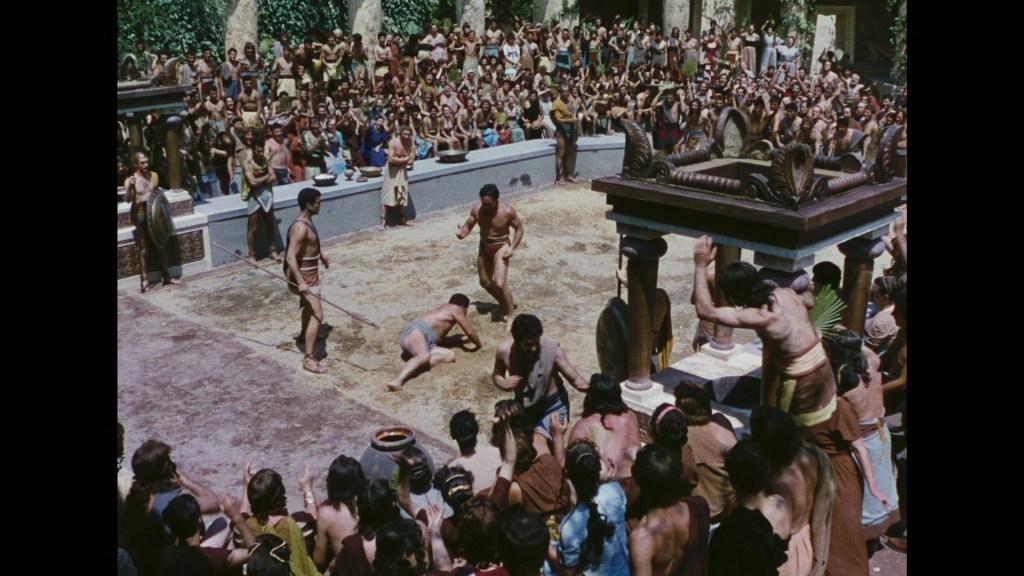 A grand arena filled with an audience watching a wrestling match, featuring several competitors, set in an ancient-themed environment.
