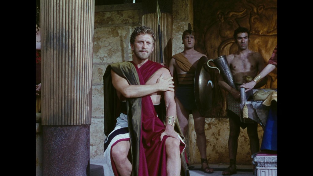 A scene featuring a man in ancient Roman attire sitting on a pedestal, flanked by two men in minimal armor and one woman dressed in colorful garments in the background.