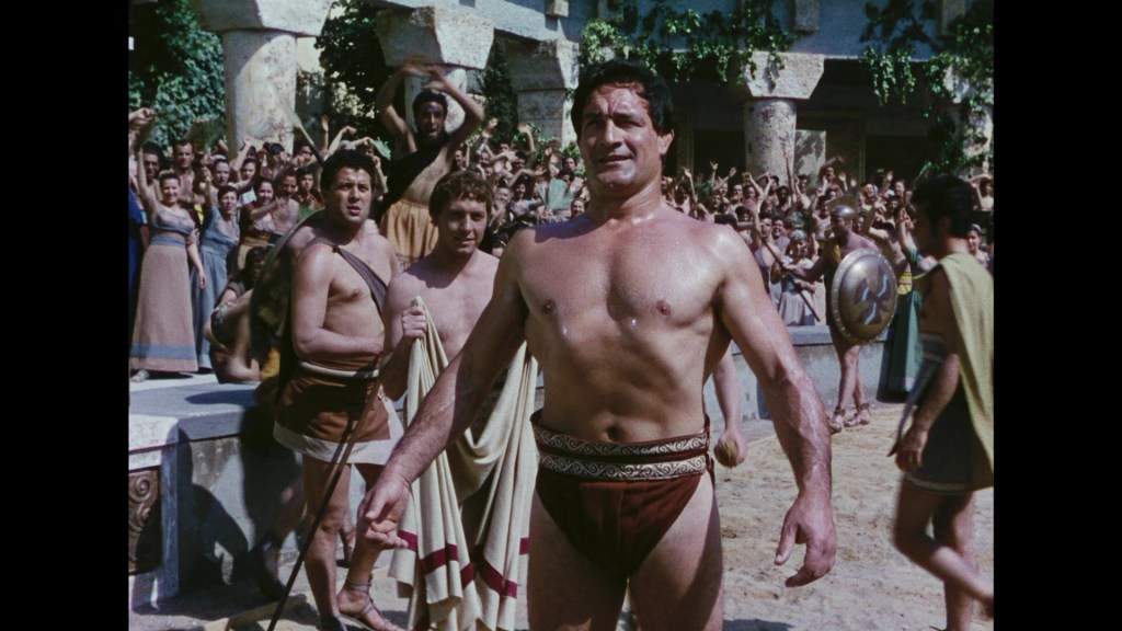 A muscular man in ancient Greek attire stands in a sandy arena, surrounded by a crowd of spectators celebrating in the background.